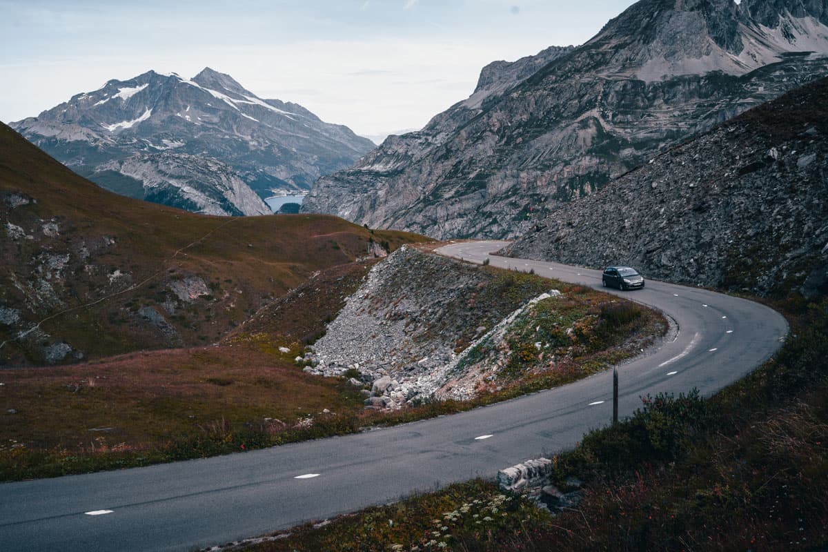 Een kronkelende bergweg slingert door rotsachtig terrein met groene hellingen, perfect voor een schilderachtige roadtrip. Een zwarte auto rijdt over de route in de Franse Alpen, met besneeuwde toppen en een blauw meer onder een bewolkte hemel.